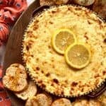 Baked ricotta cheese dip with sliced lemons on a wooden board surrounded by crostini.