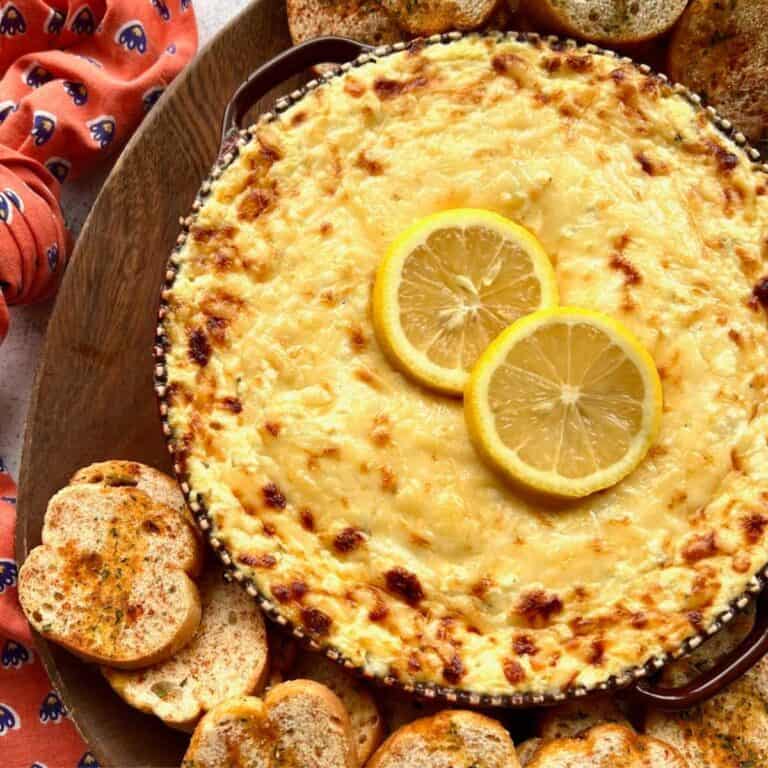 Baked ricotta cheese dip with sliced lemons on a wooden board surrounded by crostini.