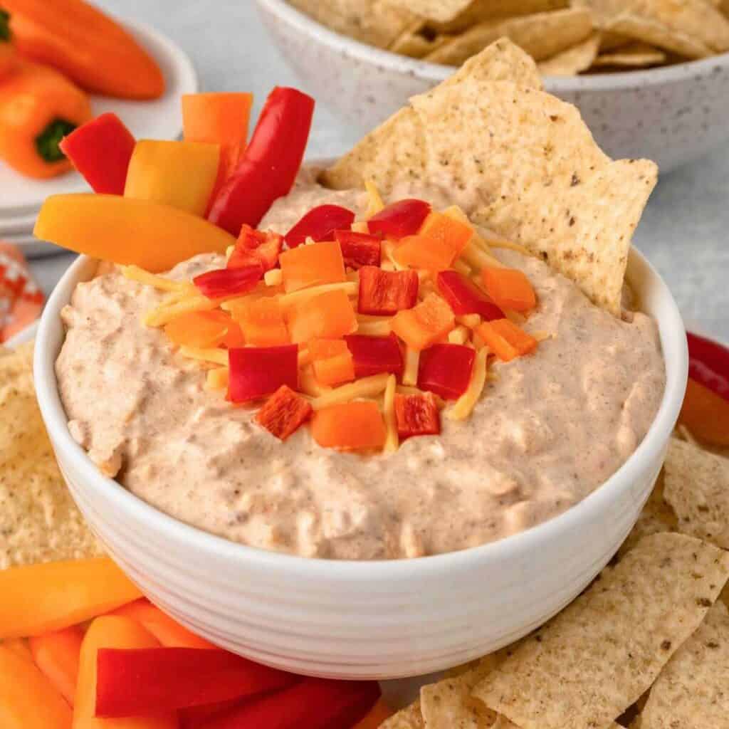 Boat dip with multi colored peppers on top in a white dish.