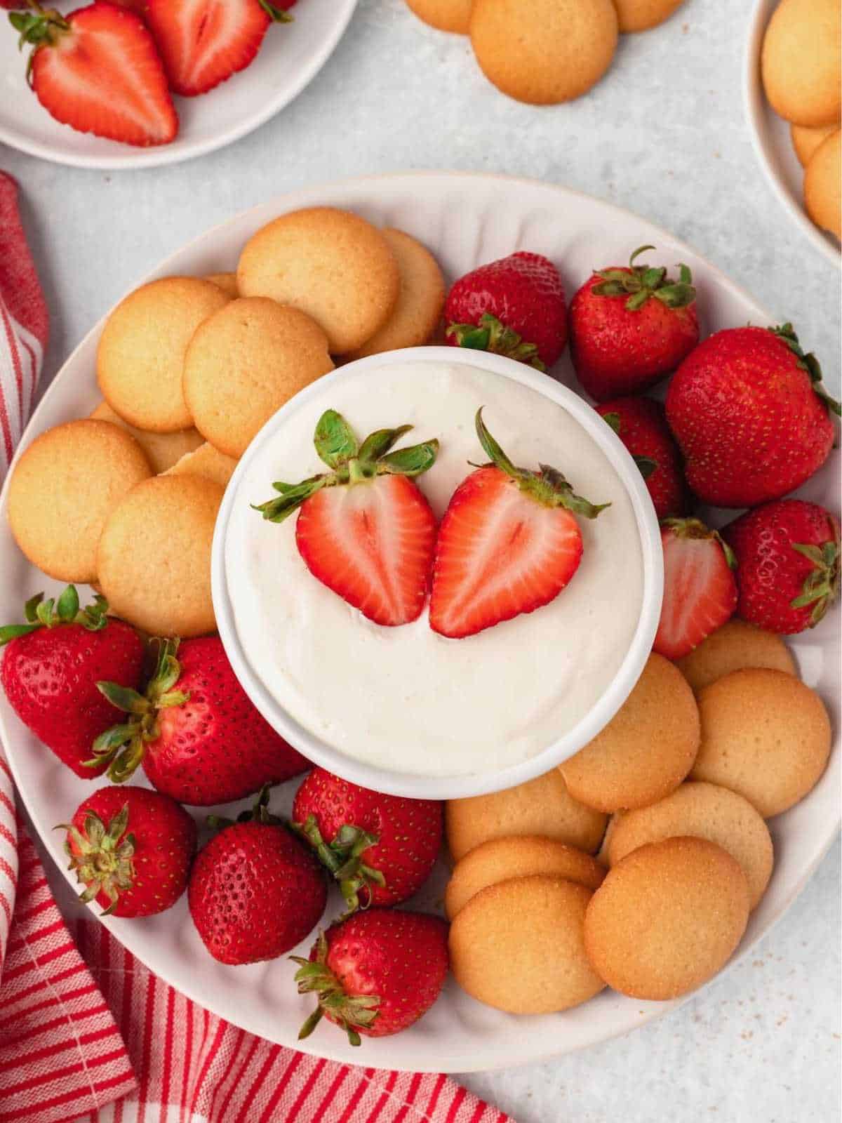 Fruit dip bowl with sliced strawberries on it on a plate of vanilla wafers and strawberries.