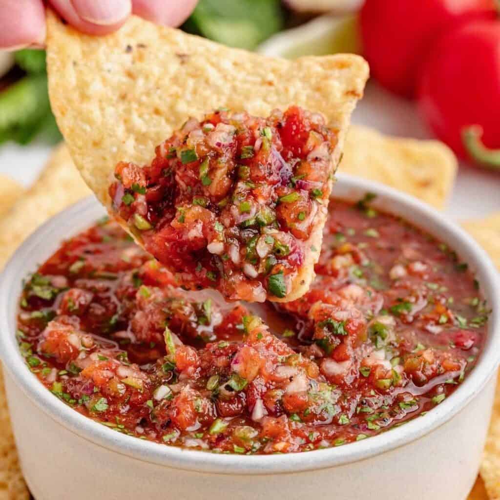 A chip being dipped into Fire roasted salsa in a white bowl.