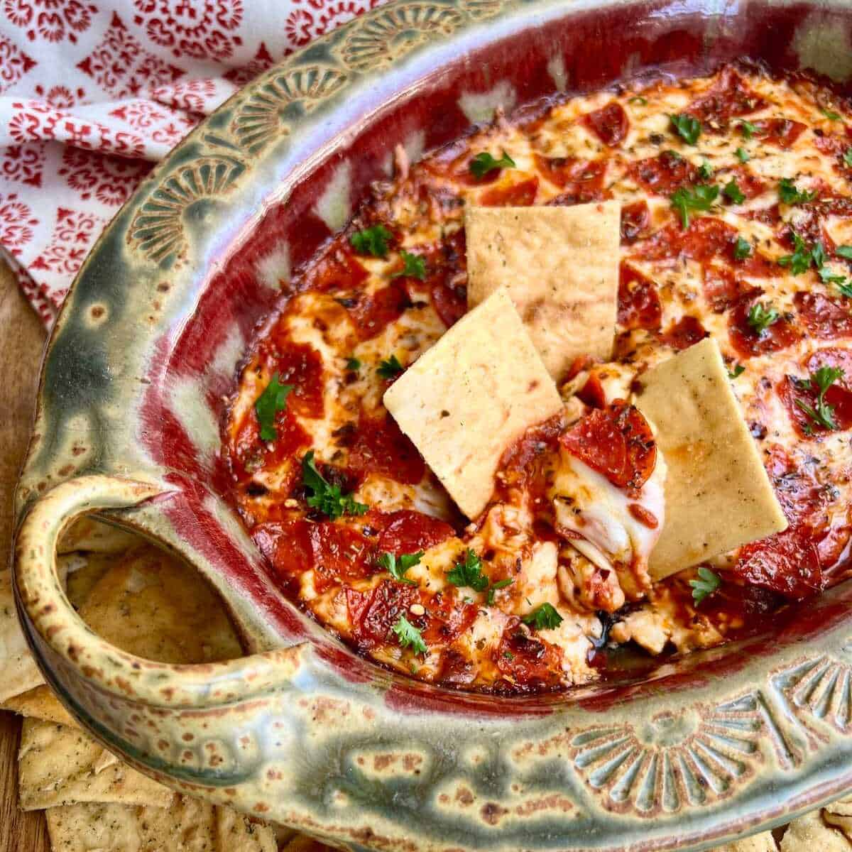 Pepperoni Pizza Dip baked in a pottery dish with crackers dipping into it.