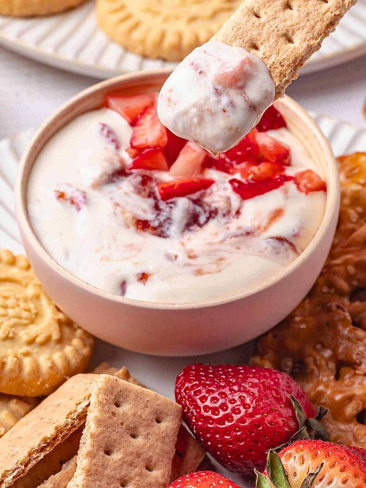 Graham cracker dipped in strawberry cheesecake dip with bowl of dip, crackers, and strawberries in the background.