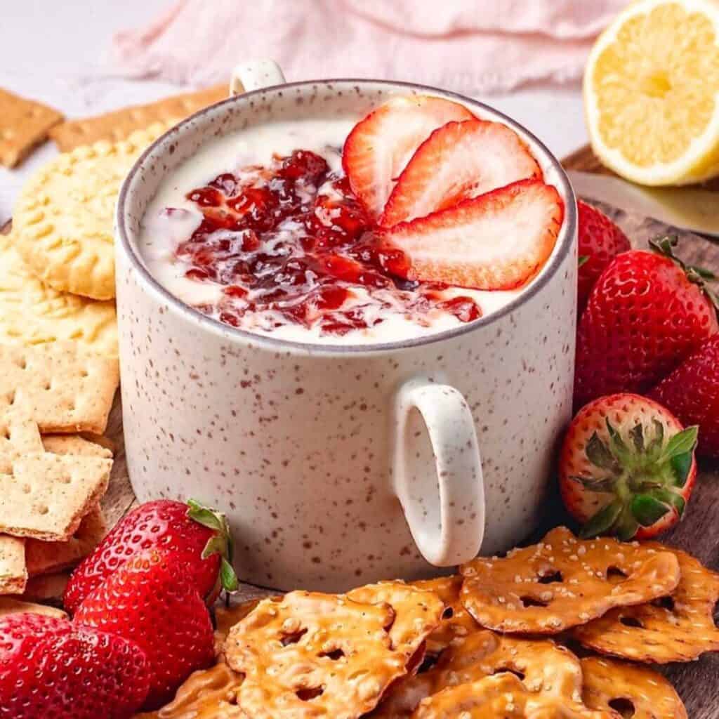 Strawberry cheesecake dip in a white mug with cookies and strawberries around it.