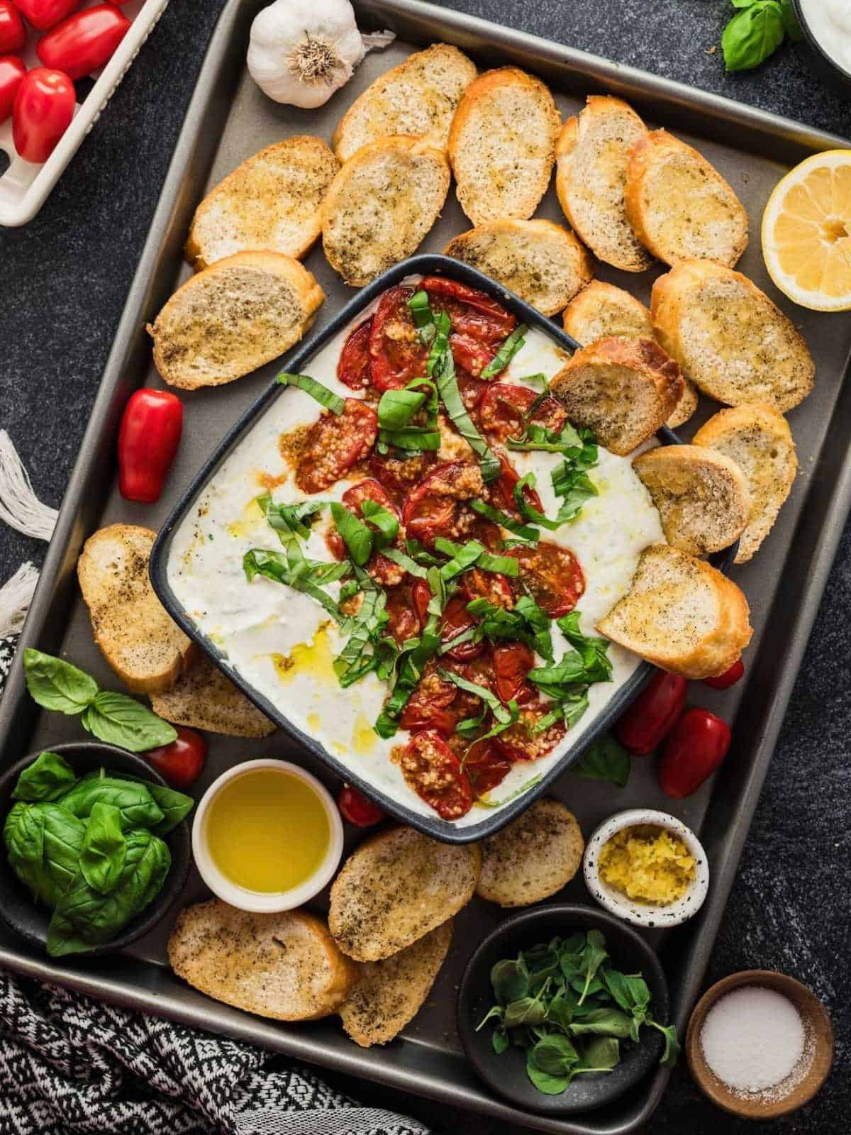 Bowl of whipped feta with roasted tomatoes dip on a plate of crostini and dip ingredients.