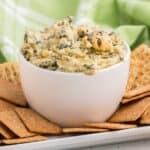 Baked spinach artichoke dip in a white bowl surrounded by crackers.