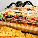 Large spider peeking over a glass dish showing 7 layer dip.