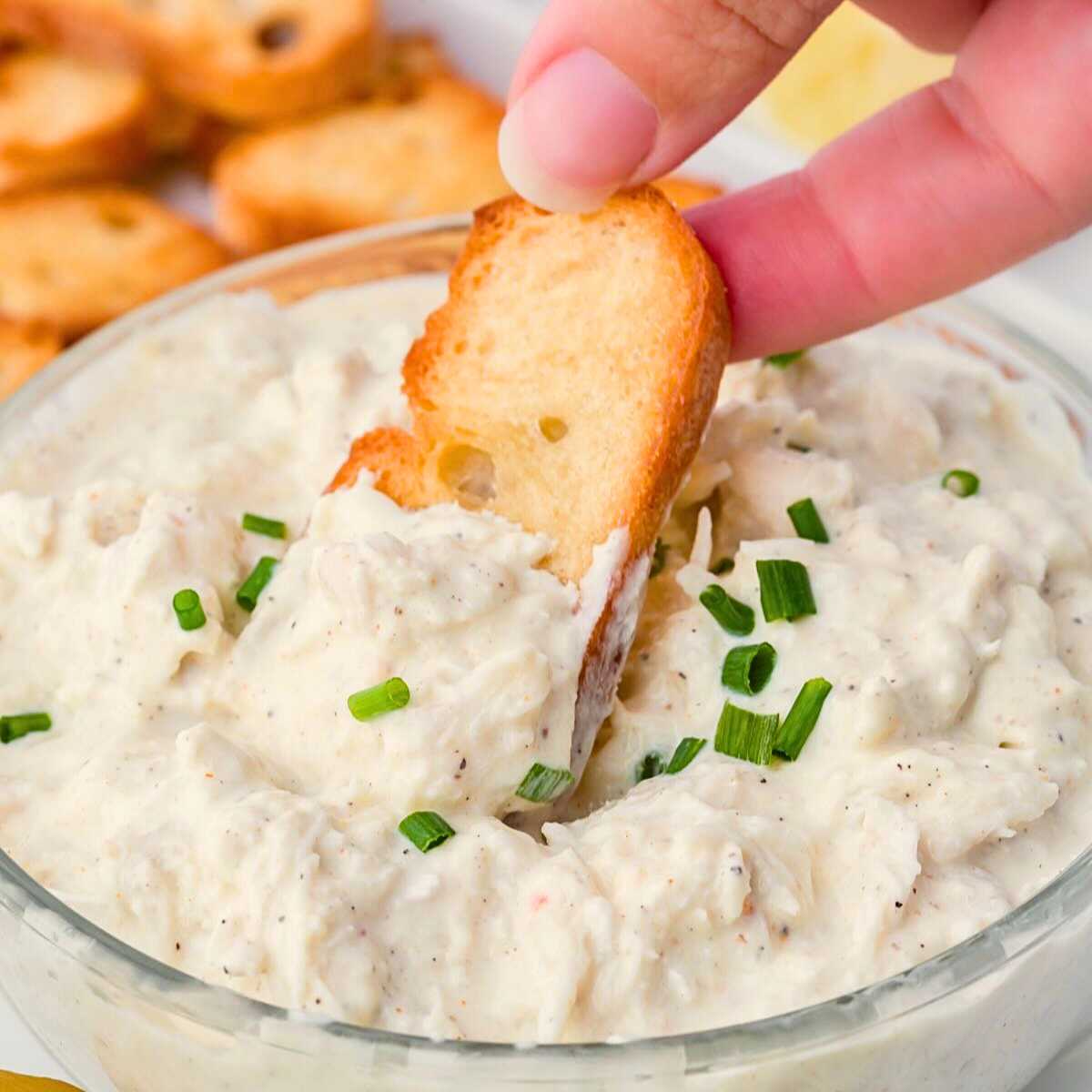 Crostini dipped into Cold Crab Dip. in a clear bowl.