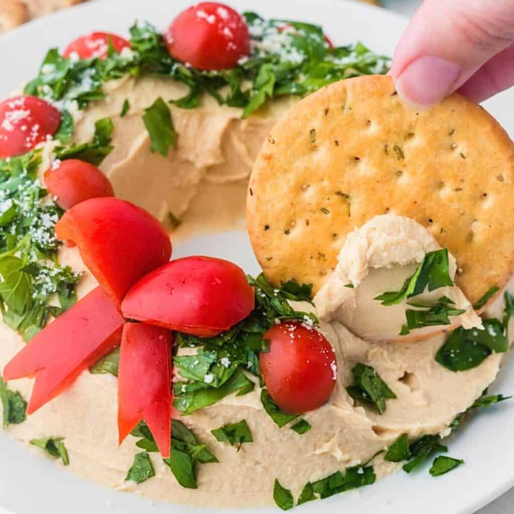 Christmas wreath made of hummus with a cracker dipping into it.