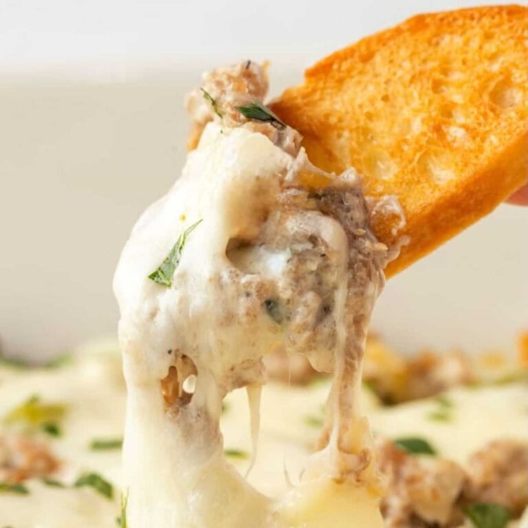 Golden crostini dipping into a bowl of Philly Cheesesteak Dip and pulling up the cheese.
