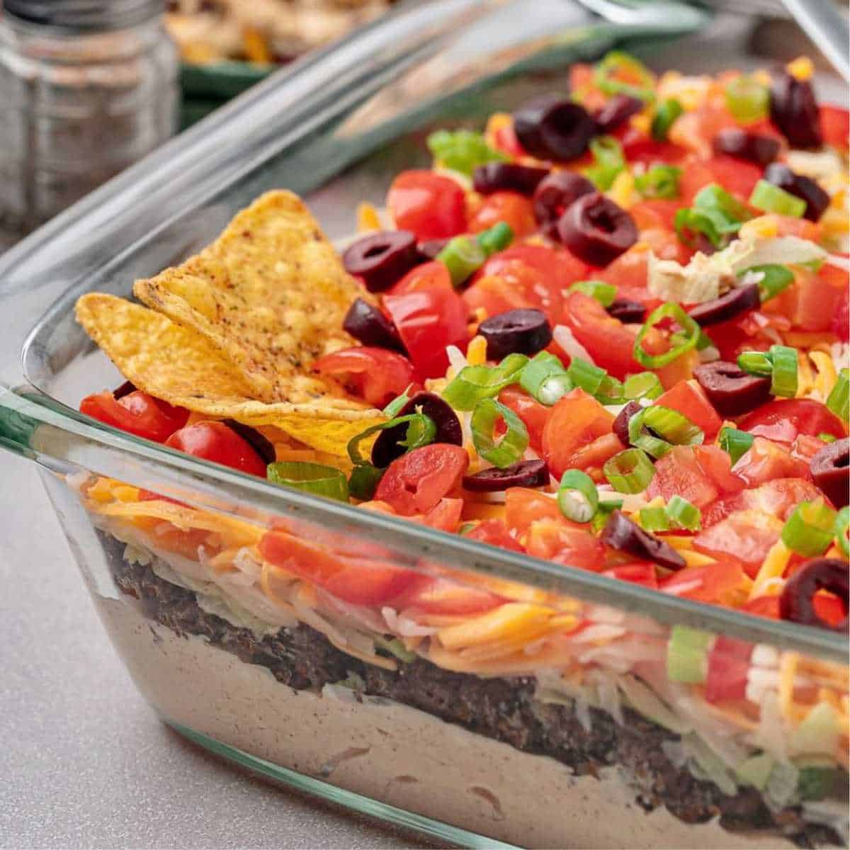 Layered Taco Dip in a clear dish with tortilla chips.