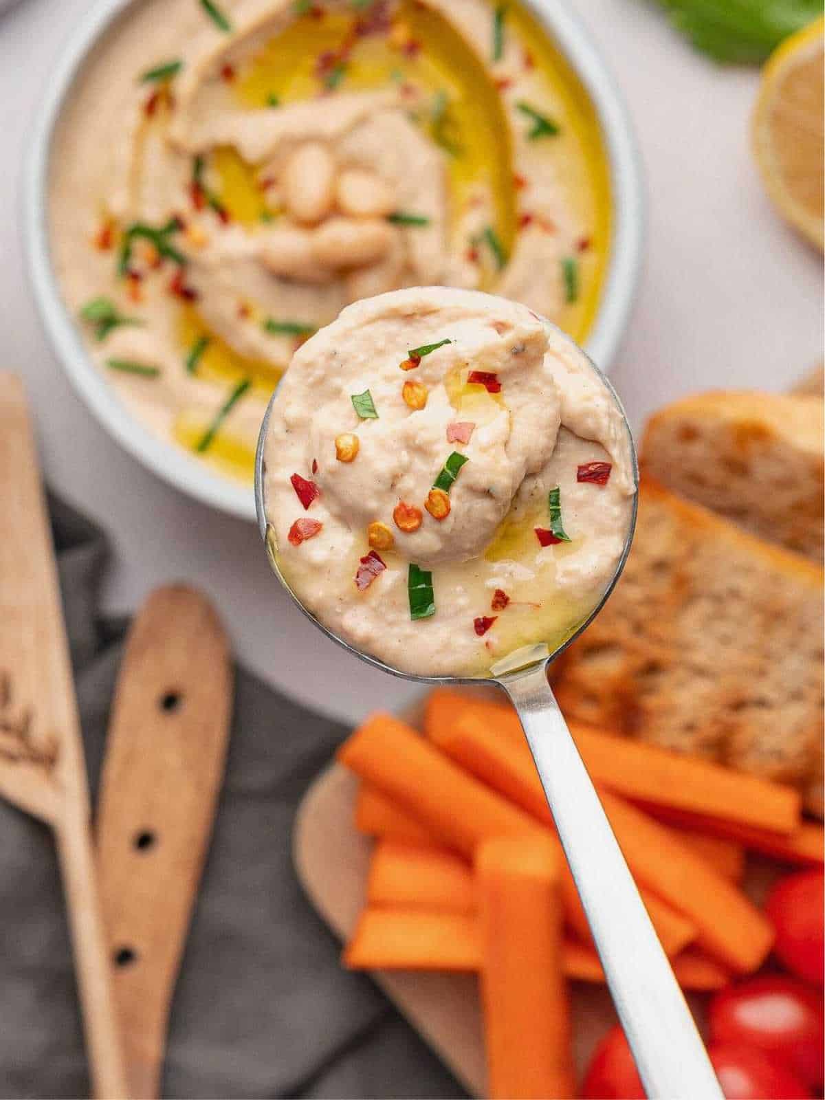 A scoop of white bean dip held up over a bowl with veggies surrounding it.