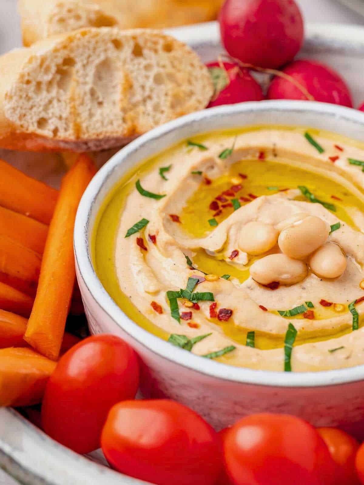 A white bowl filled with cannellini bean dip, tomatoes, carrots and radishes.