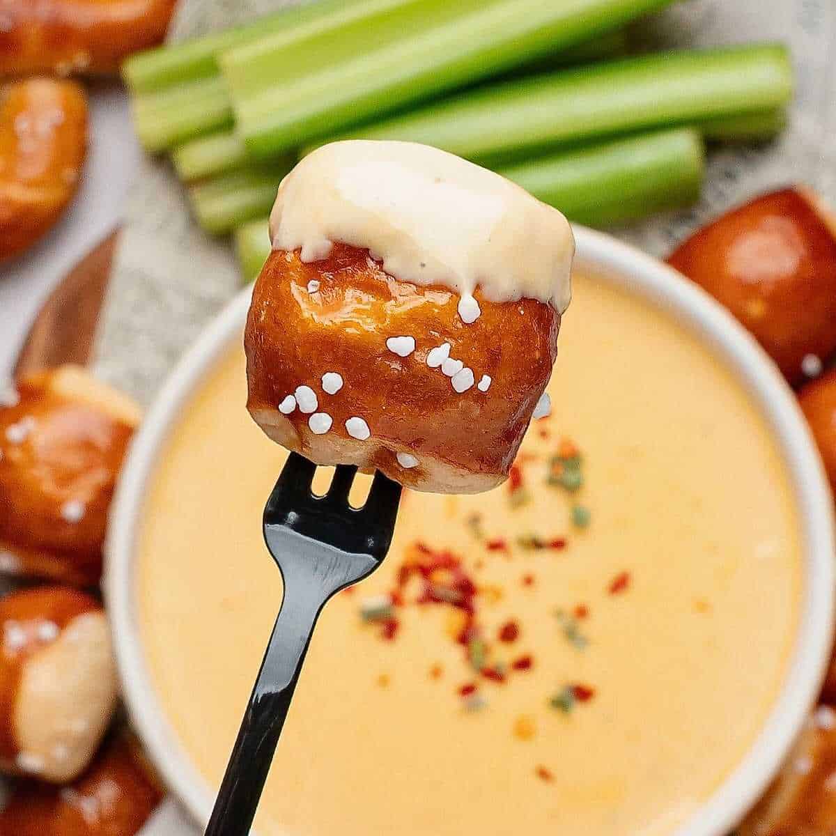 Beer cheese pretzel dip being lifted out of a bowl by a pretzel bite on a fork.