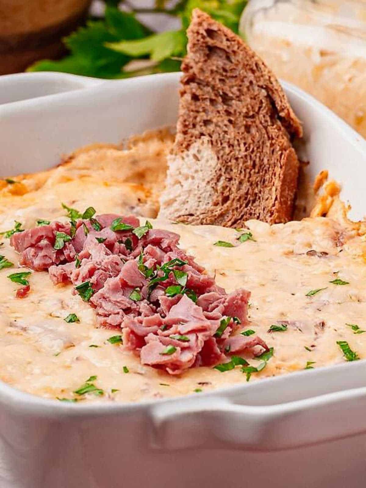 Rye bread dipping into cheesy reuben dip in a white baking dish.