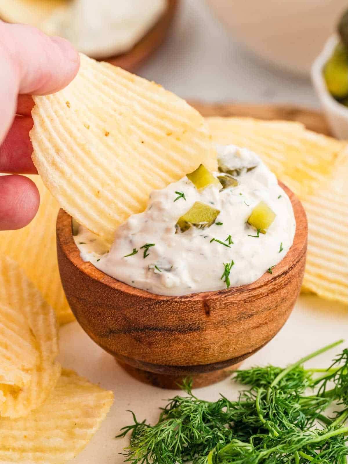 Potato chip dipping into a wooden bowl of dill pickle dip.