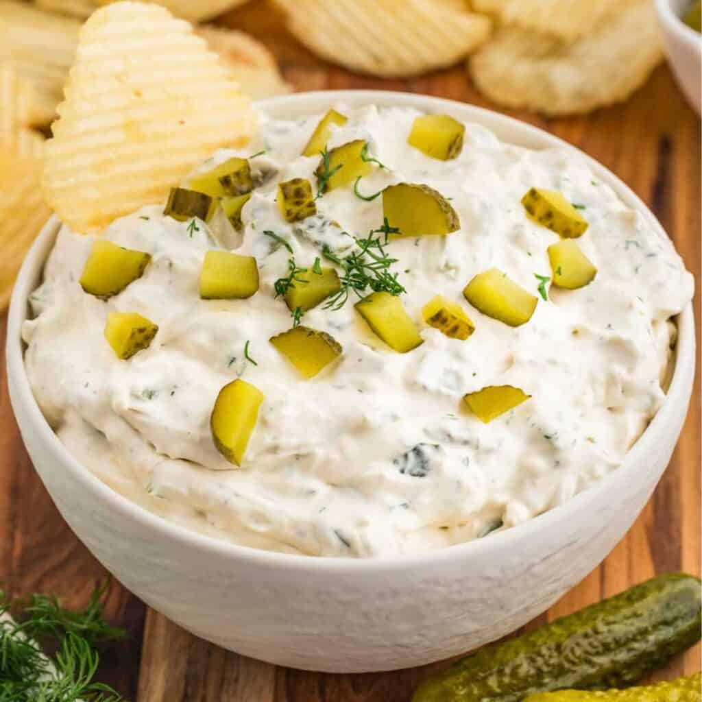 Dill pickle dip with pickle chips on top and a potato chip dipping into a bowl.