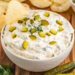 Dill pickle dip with pickle chips on top and a potato chip dipping into a bowl.