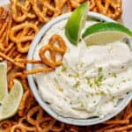 Bowl of Creamy Margarita Dip with 2 lime wedges surrounded by salted pretzels.