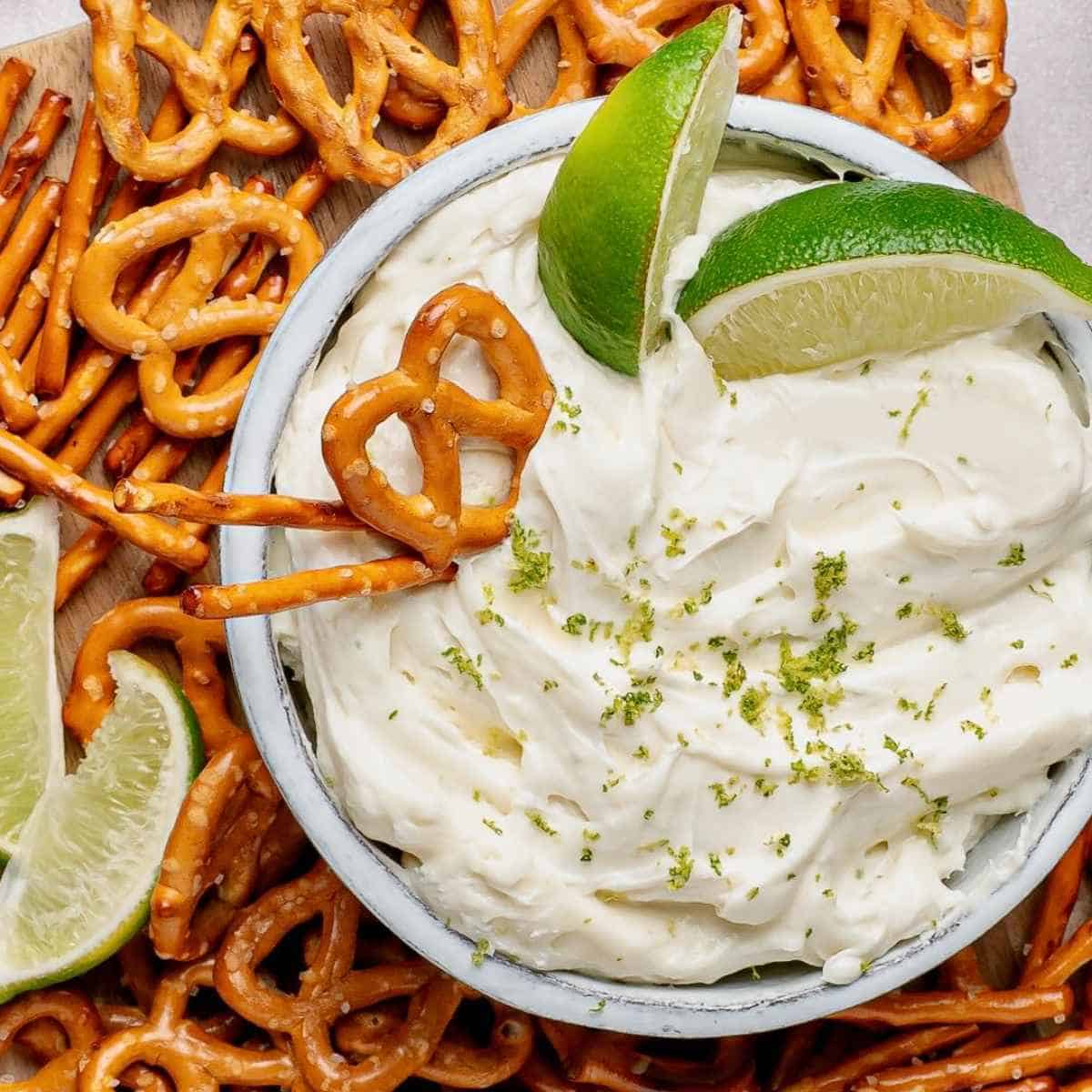 Bowl of Creamy Margarita Dip with 2 lime wedges surrounded by salted pretzels.