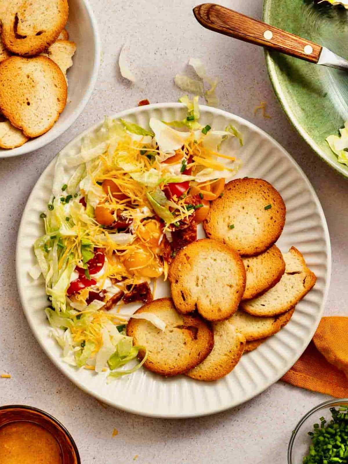 Lettuce, tomatoes and cheese on top of cream cheese mixture with bagel chips on a white plate.