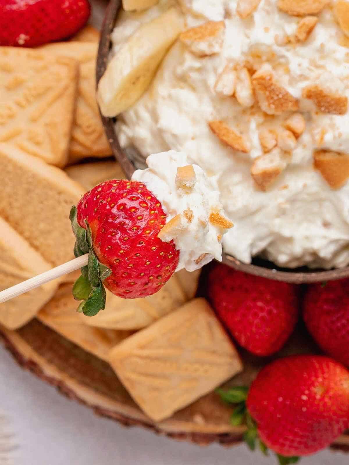 A strawberry on a stick dipping into banana pudding dip.