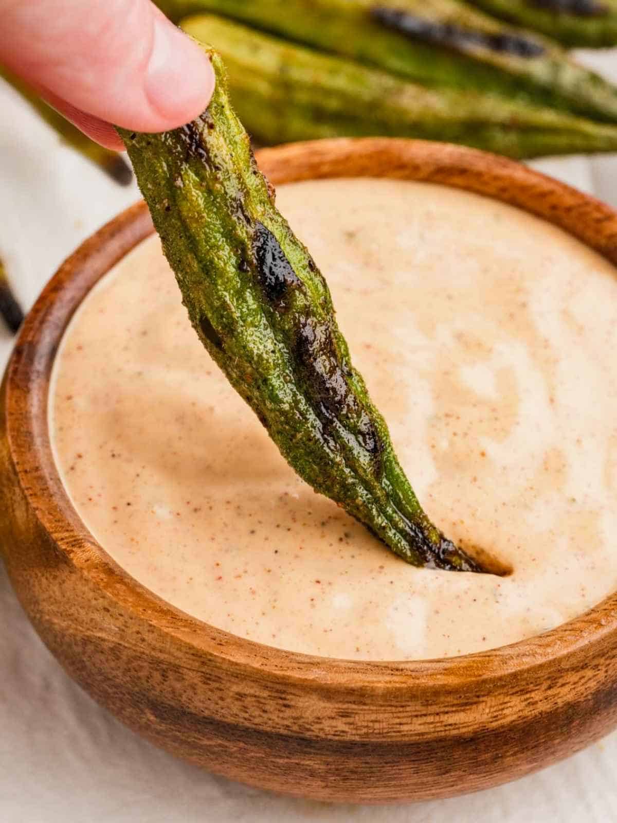 Grilled okra being dipped into a brown bowl of Cajun dipping sauce.