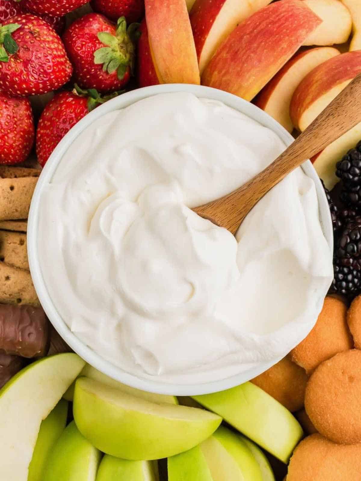 White bowl of fruit dip with a wooden spoon surrounded by fruit.