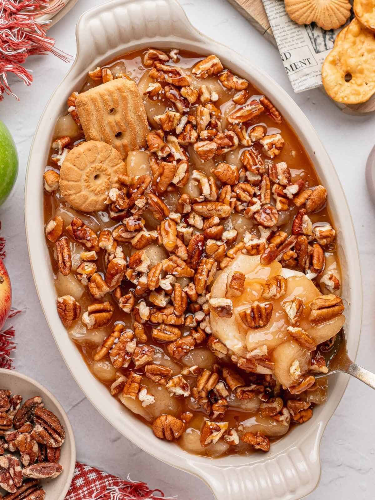 Spoon of caramel apple dip being lifted up over pecans and caramel in dish.