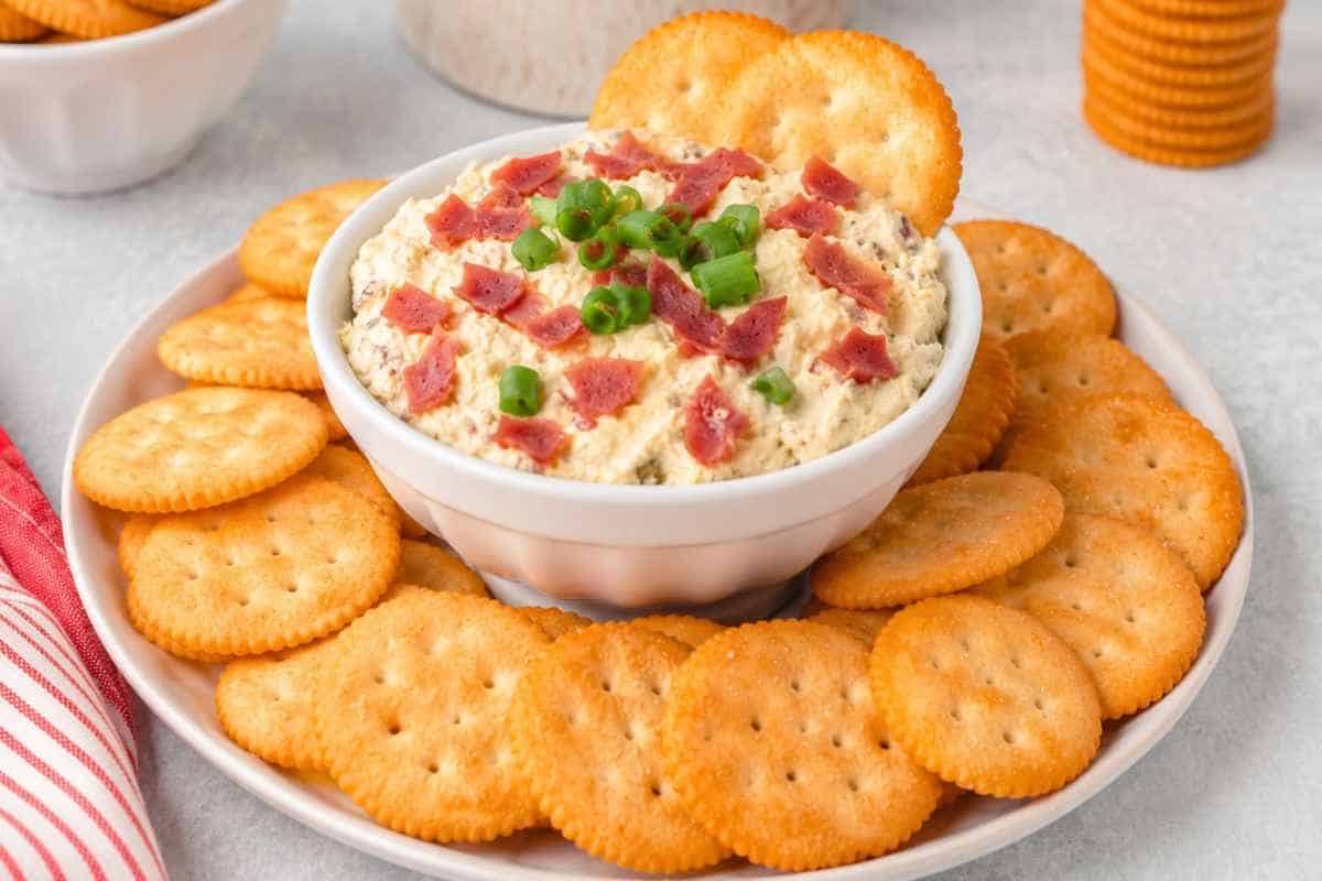 Hot Chipped Beef Dip in a white bowl surrounded by Ritz Crackers topped with onions.