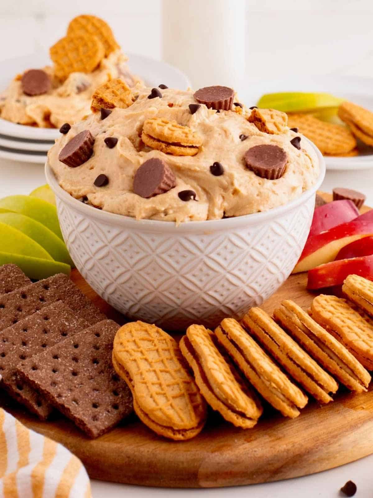 Large bowl of peanut butter dip surrounded by cookies and apple slices.