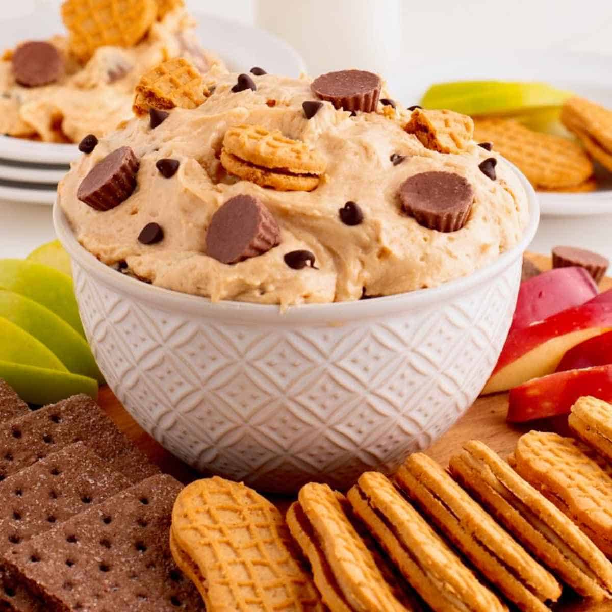 Peanut butter dip topped with Reese's cups and cookies on top surrounded by apple slices and cookies.