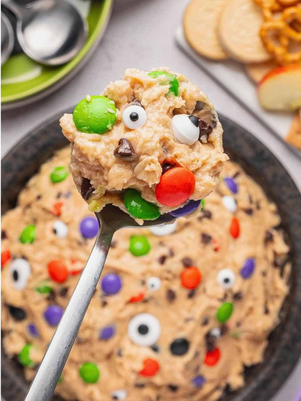 Large spoon holding up a scoop of edible cookie dough with candy eyes and Halloween M&Ms.