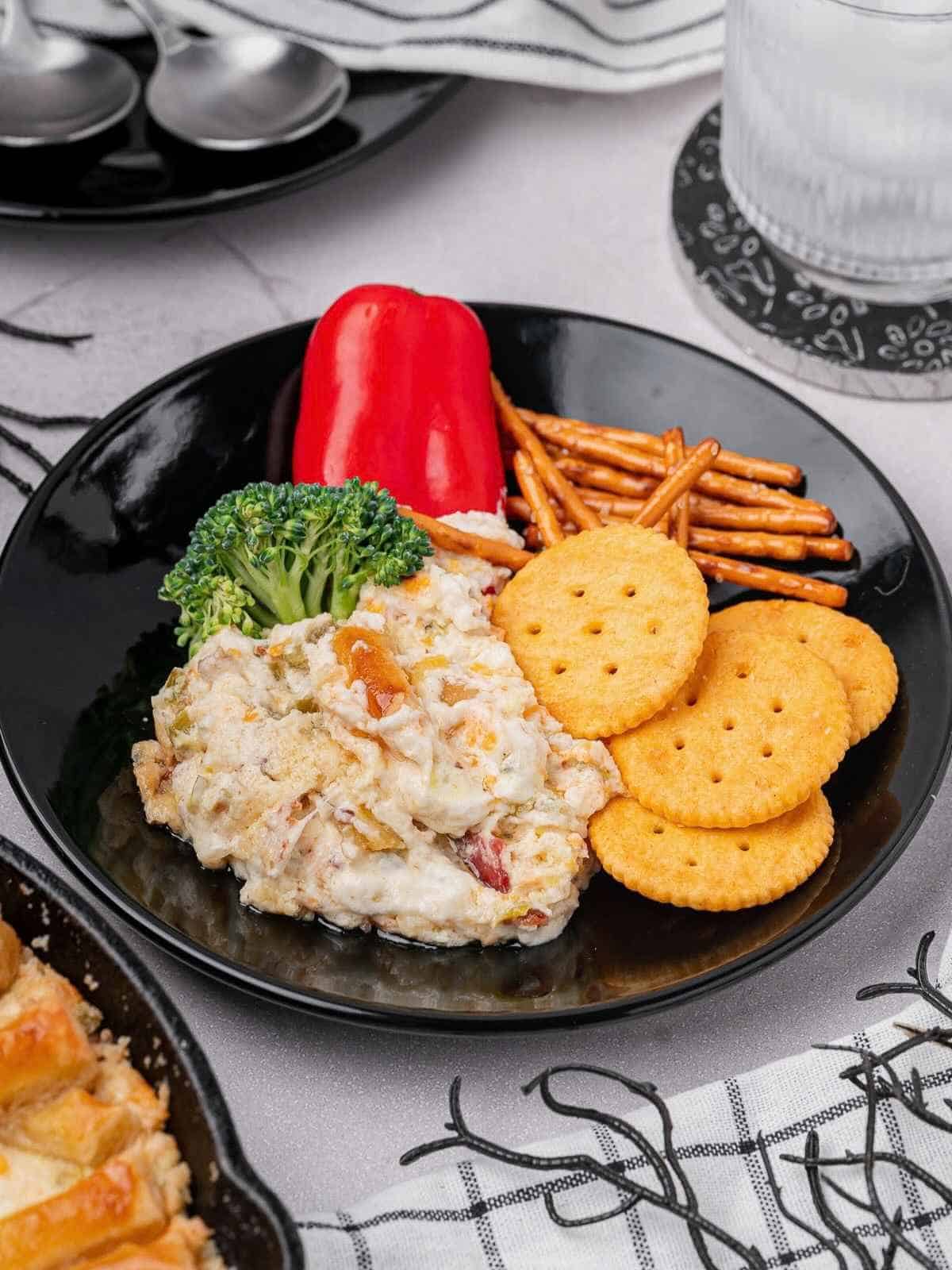 Black plate with crackers, a red pepper, broccoli and pretzels dipping into jalapeno popper dip.