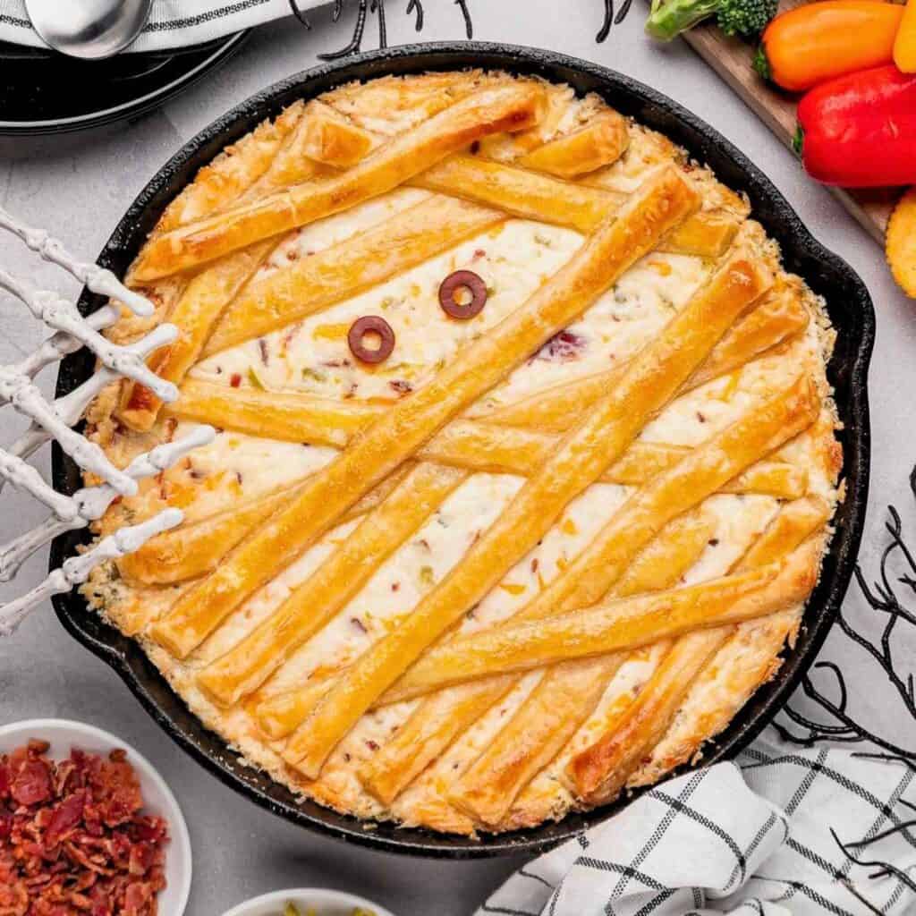 Jalapeno popper dip with mummy bandages and olive eyes in a cast iron skillet.